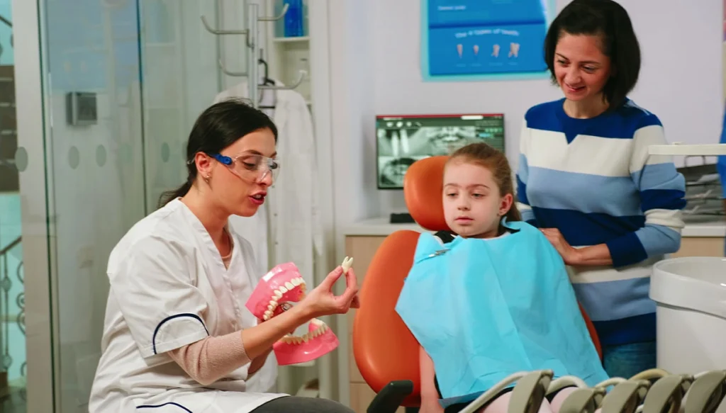 dentist speaks with child while holding a model of dentures while parent watches